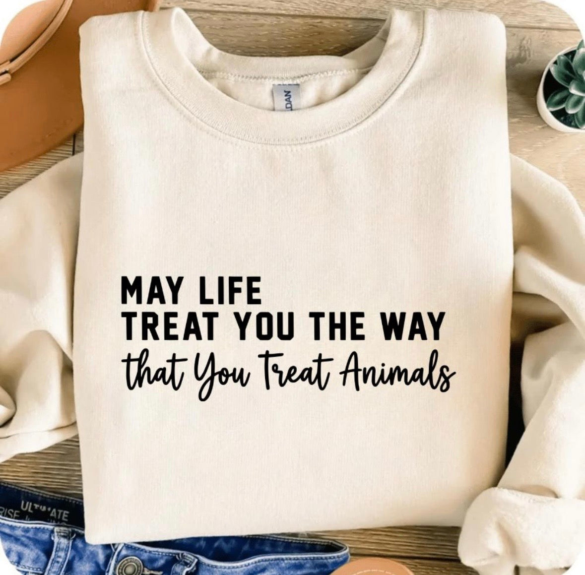 A folded cream sweatshirt with black printed text reading 'MAY LIFE TREAT YOU THE WAY that You Treat Animals' on a wooden surface, styled with a hat, jeans, and a small plant.