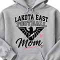 A close-up of a light heather gray hoodie featuring a graphic print that reads 'LAKOTA EAST FOOTBALL Mom' with a black eagle logo, lightning bolt, and pink hearts.