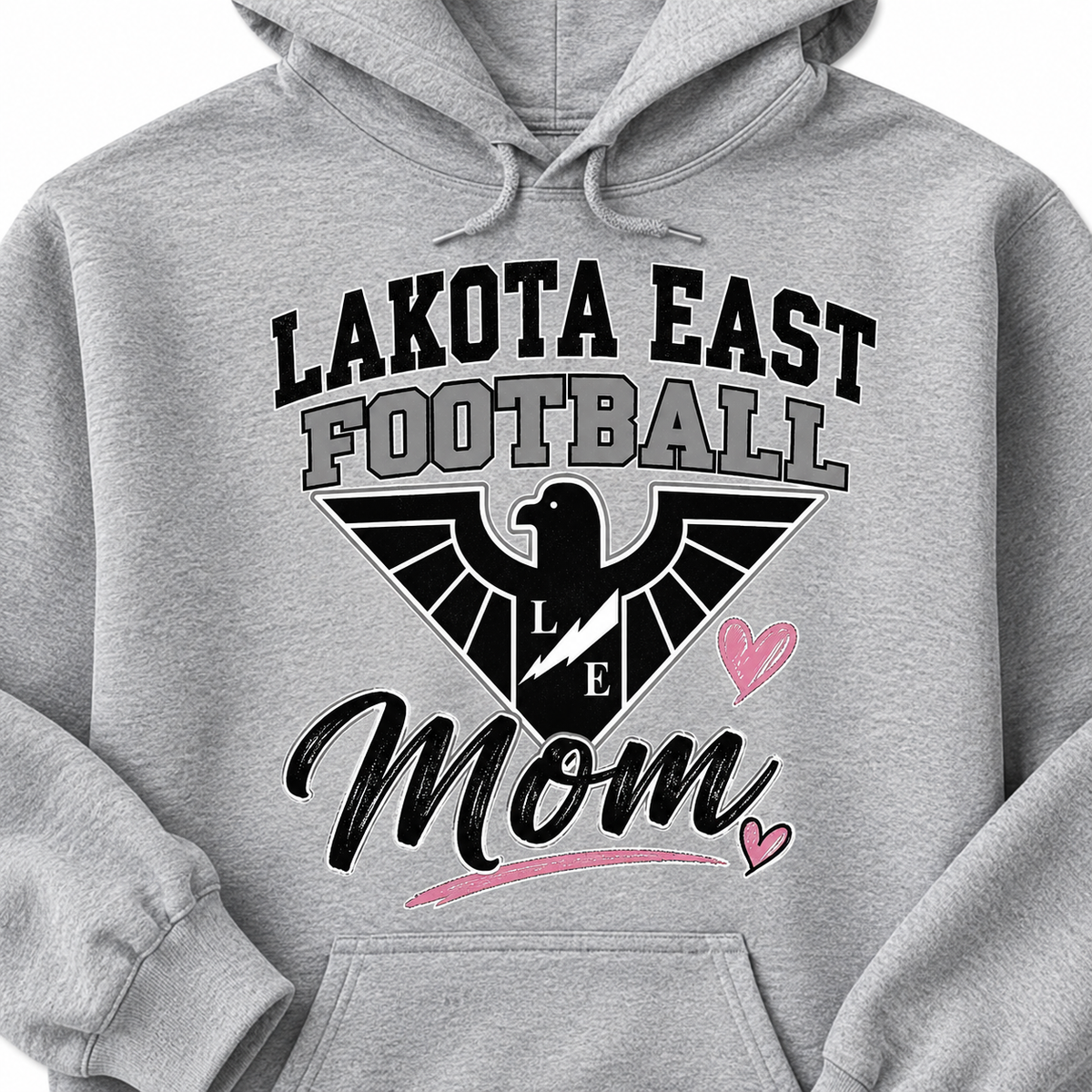 A close-up of a light heather gray hoodie featuring a graphic print that reads 'LAKOTA EAST FOOTBALL Mom' with a black eagle logo, lightning bolt, and pink hearts.