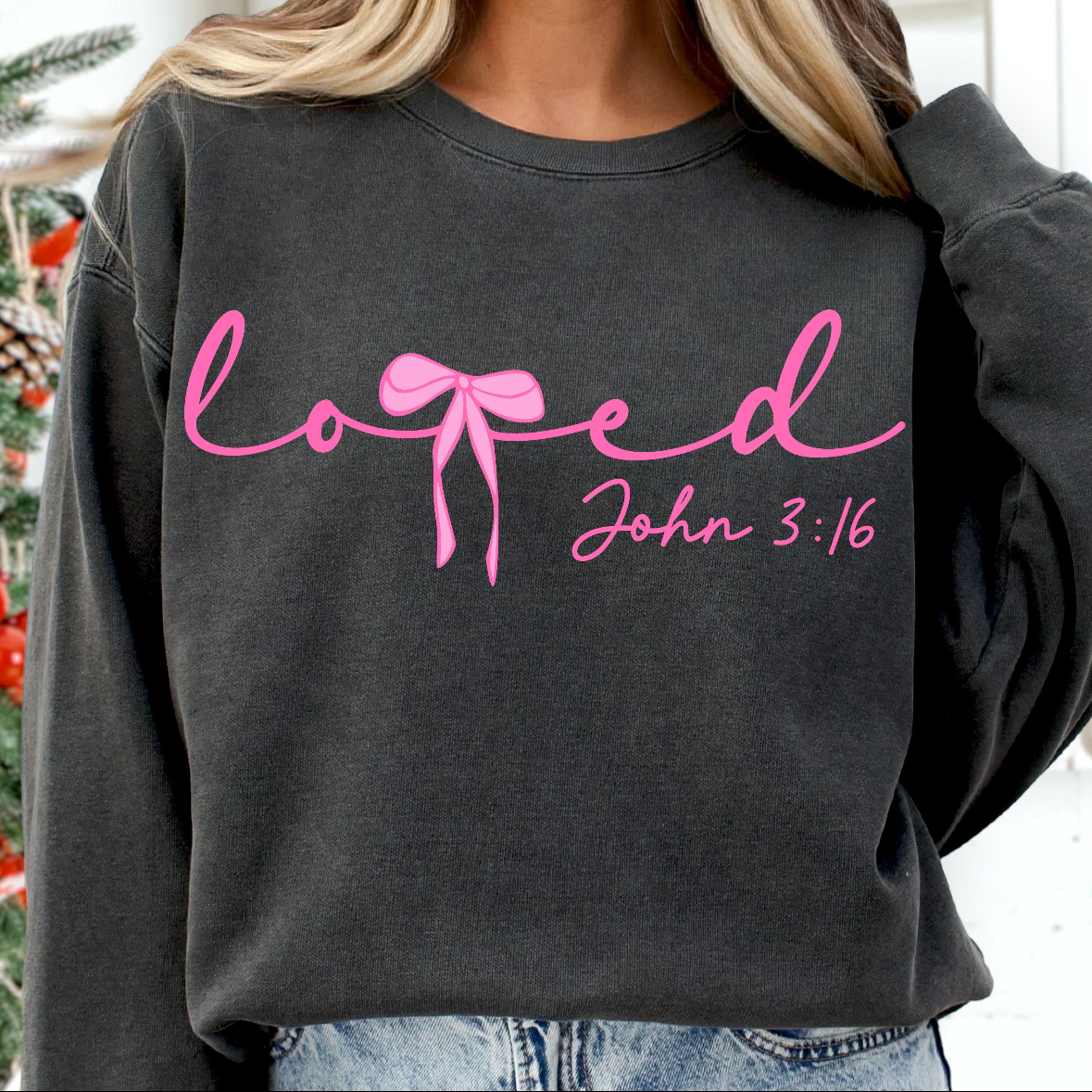 A person wearing a dark charcoal gray sweatshirt with pink script text reading 'loved' and 'John 3:16' across the chest, paired with light blue jeans. The photo is cropped from the neck down, with black bars and an Apple Pay payment banner visible around it.