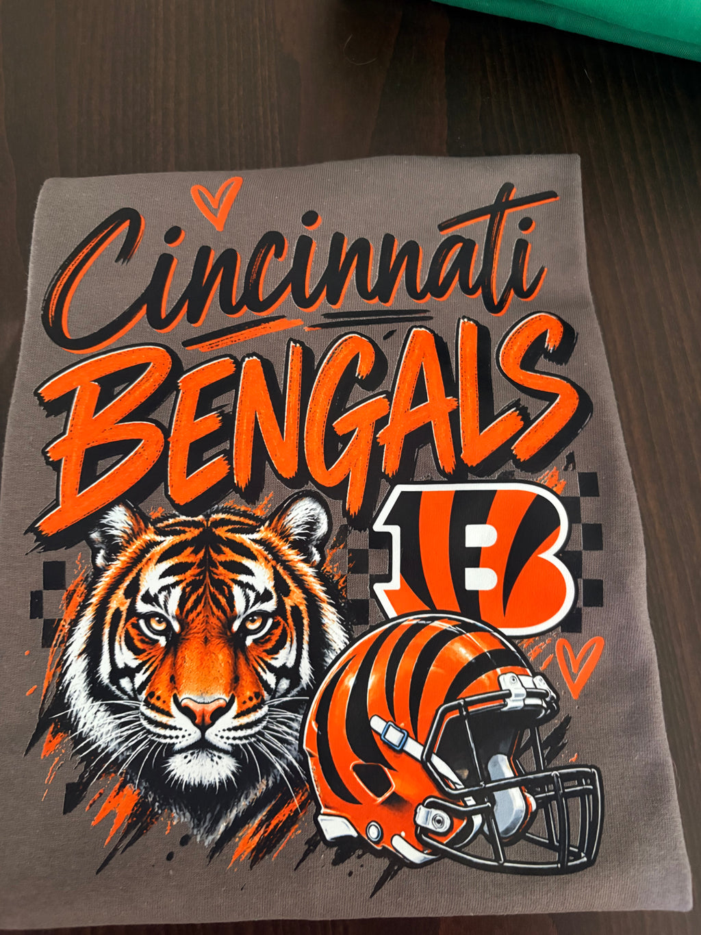 A gray graphic t-shirt featuring the Cincinnati Bengals logo, a tiger illustration, and a football helmet, laid flat on a dark wooden surface.