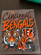 A gray graphic t-shirt featuring the Cincinnati Bengals logo, a tiger illustration, and a football helmet, laid flat on a dark wooden surface.