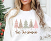 White sweatshirt with Christmas tree design and 'Tis The Season text, worn by a person in front of a decorated Christmas tree.