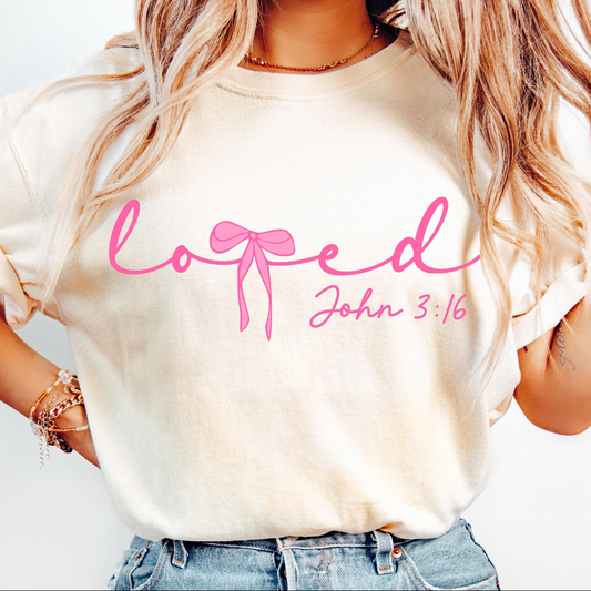 A woman wearing a cream-colored t-shirt with pink text that reads 'loved John 3:16' with a pink bow graphic, paired with blue jeans.
